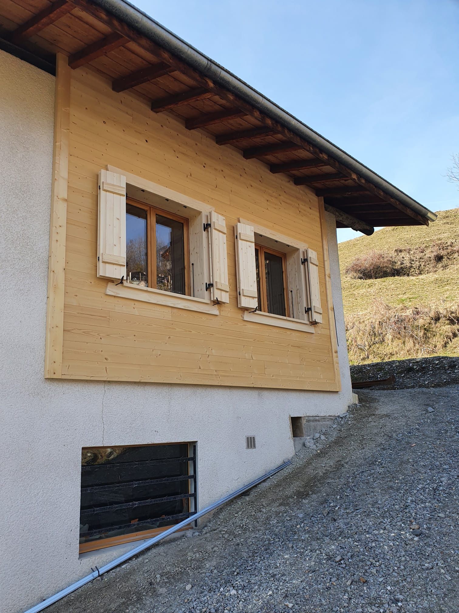 Petite maison beige aux volets en bois, située au bord d'une allée de gravier en pente, vue d'en bas.