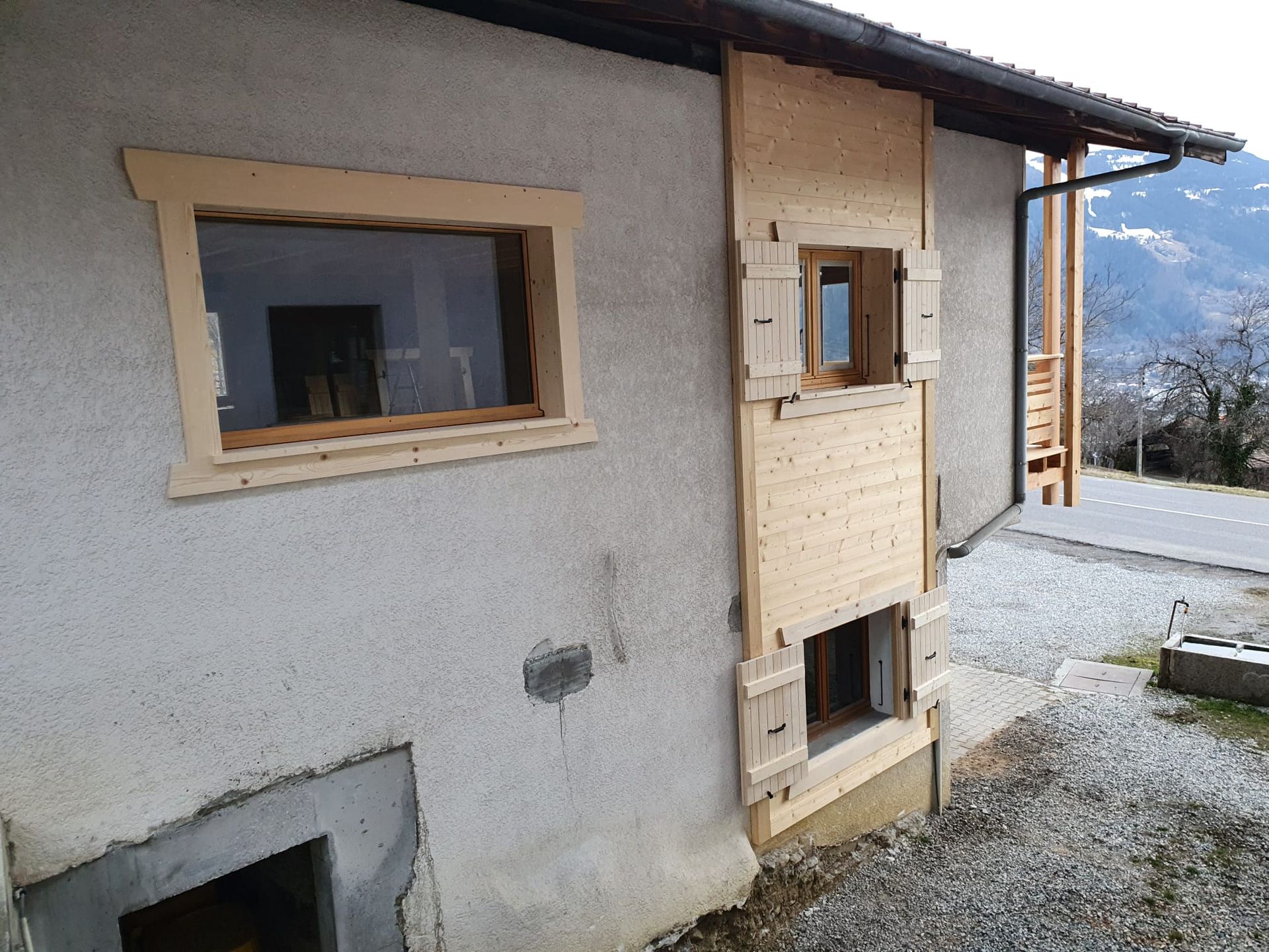 Façade de maison patinée par le temps, fenêtres condamnées et allée de gravier, sur fond de paysage montagneux.