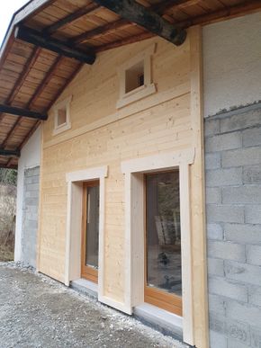 Extérieur d'une petite maison inachevée, en construction, avec deux hautes fenêtres et un auvent en bois.