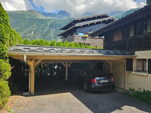 Un abri de voiture avec une voiture noire garée à côté d'un chalet, donnant sur des montagnes verdoyantes sous un ciel nuageux.