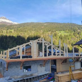Charpente de toit en bois partiellement construite, avec montagnes et forêt en arrière-plan.