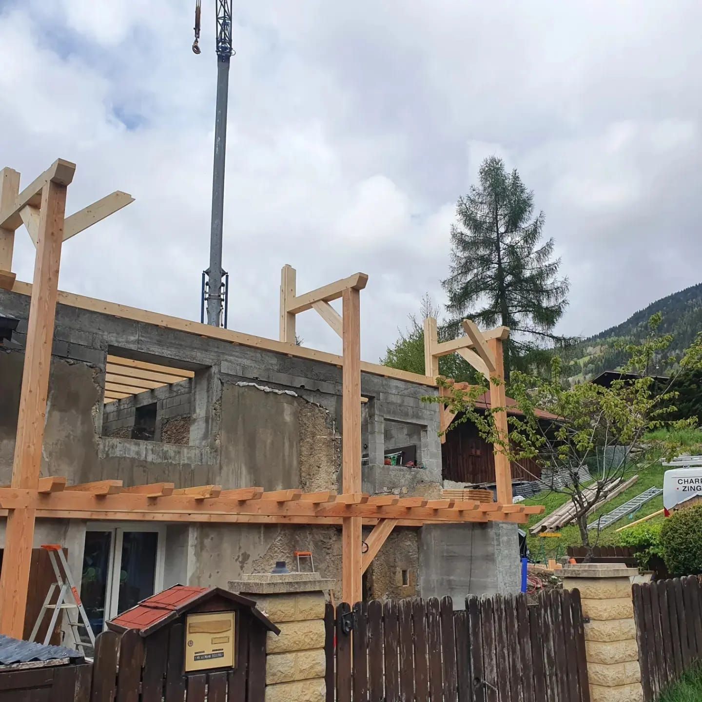Maison en béton partiellement construite avec ossature en bois, cour clôturée et vue sur une montagne nuageuse en arrière-plan.