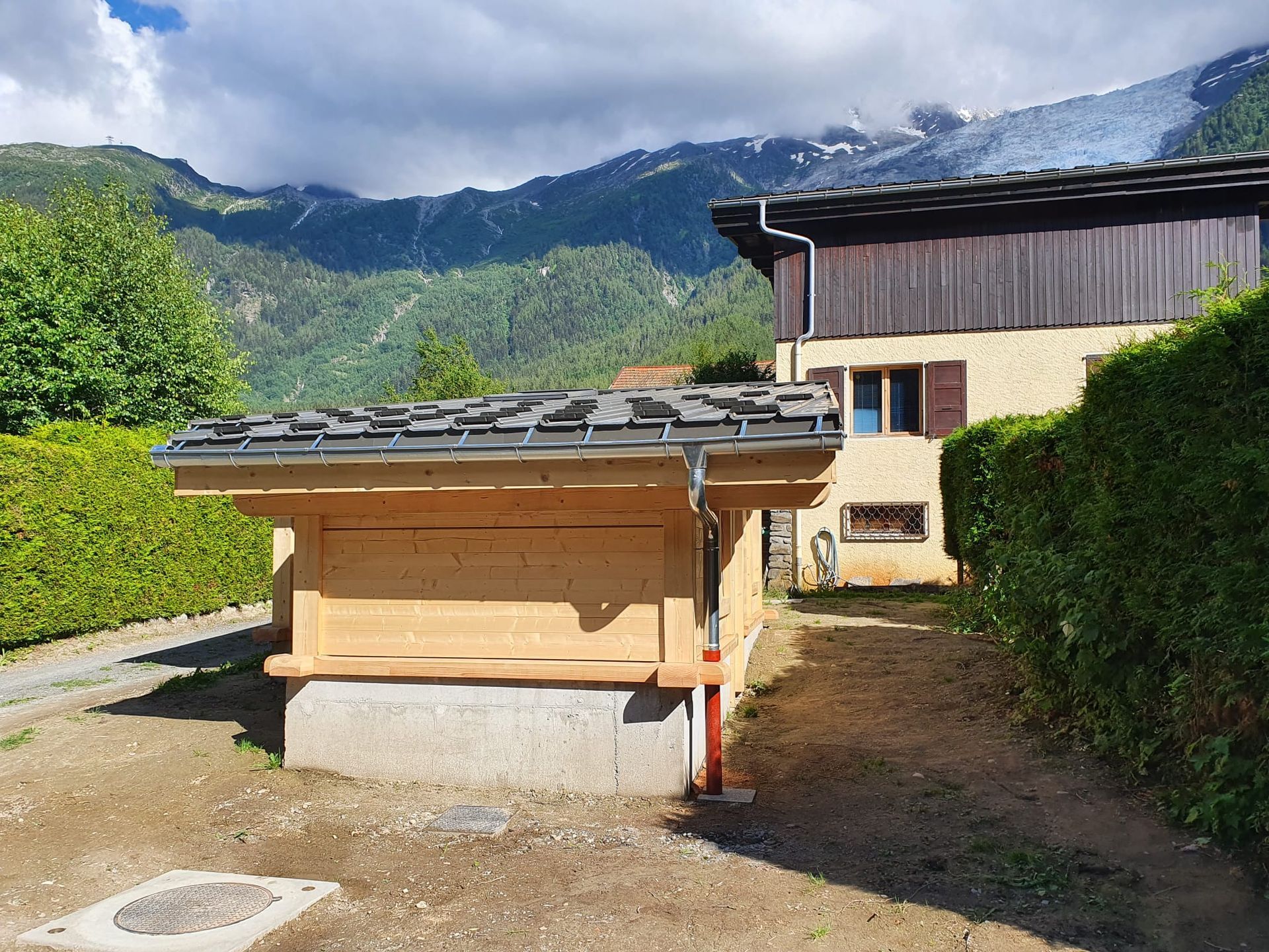 Structure en bois à côté d'une maison, avec des montagnes et des nuages en arrière-plan.