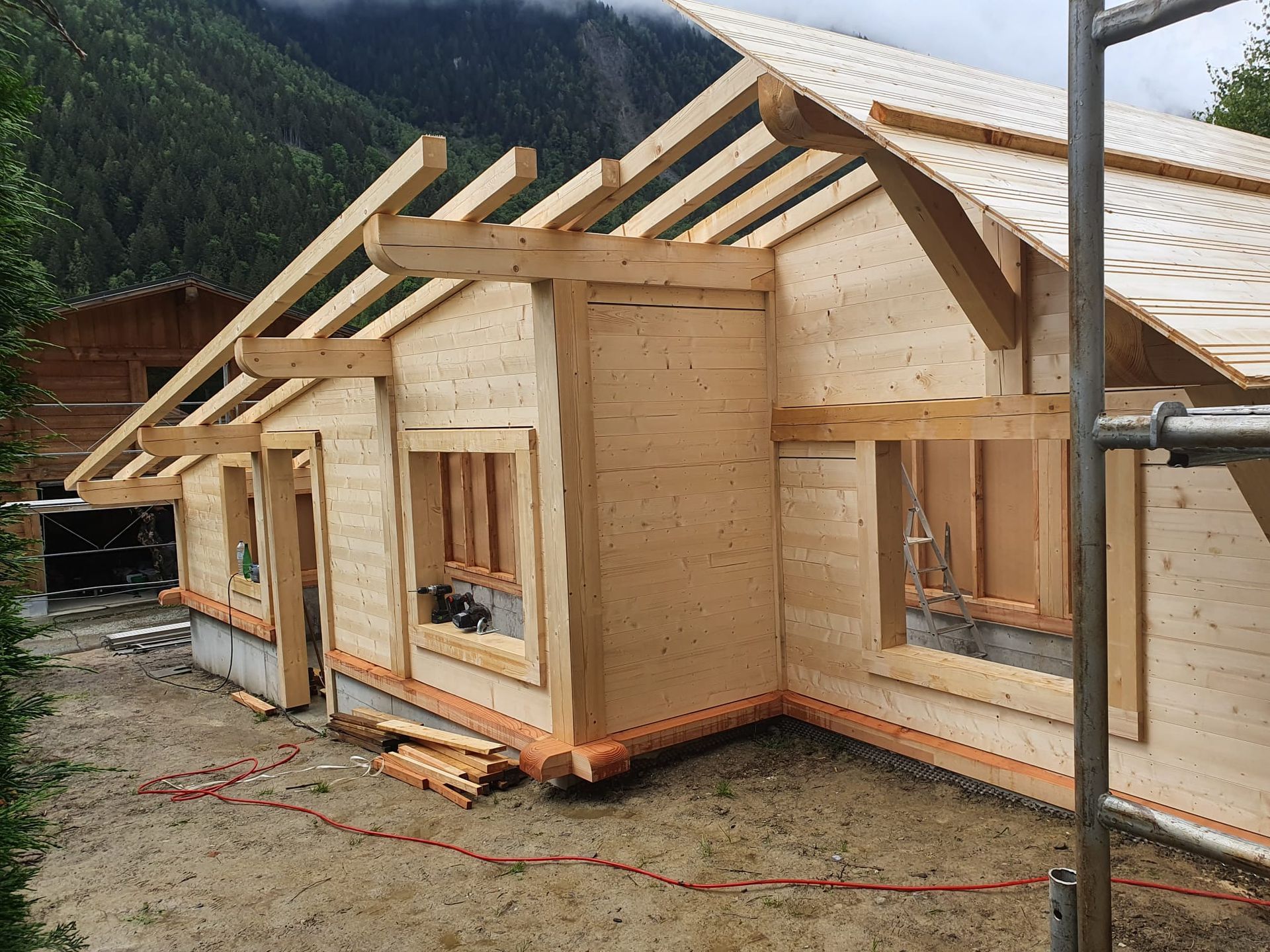 Maison à ossature en bois en construction avec charpente apparente et ouvertures de fenêtres sur un terrain boueux