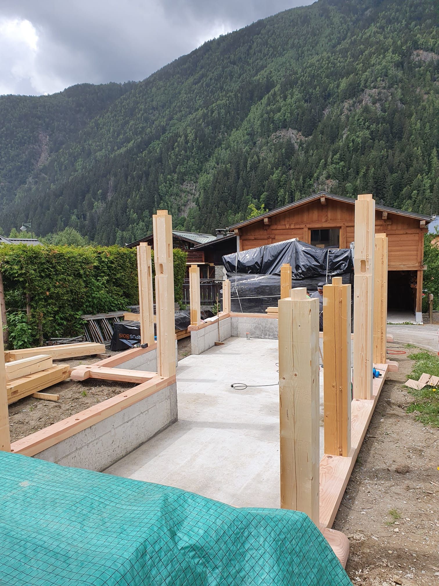 Allée en béton partiellement construite avec des poteaux en bois, se détachant sur fond de montagnes verdoyantes et de ciel nuageux