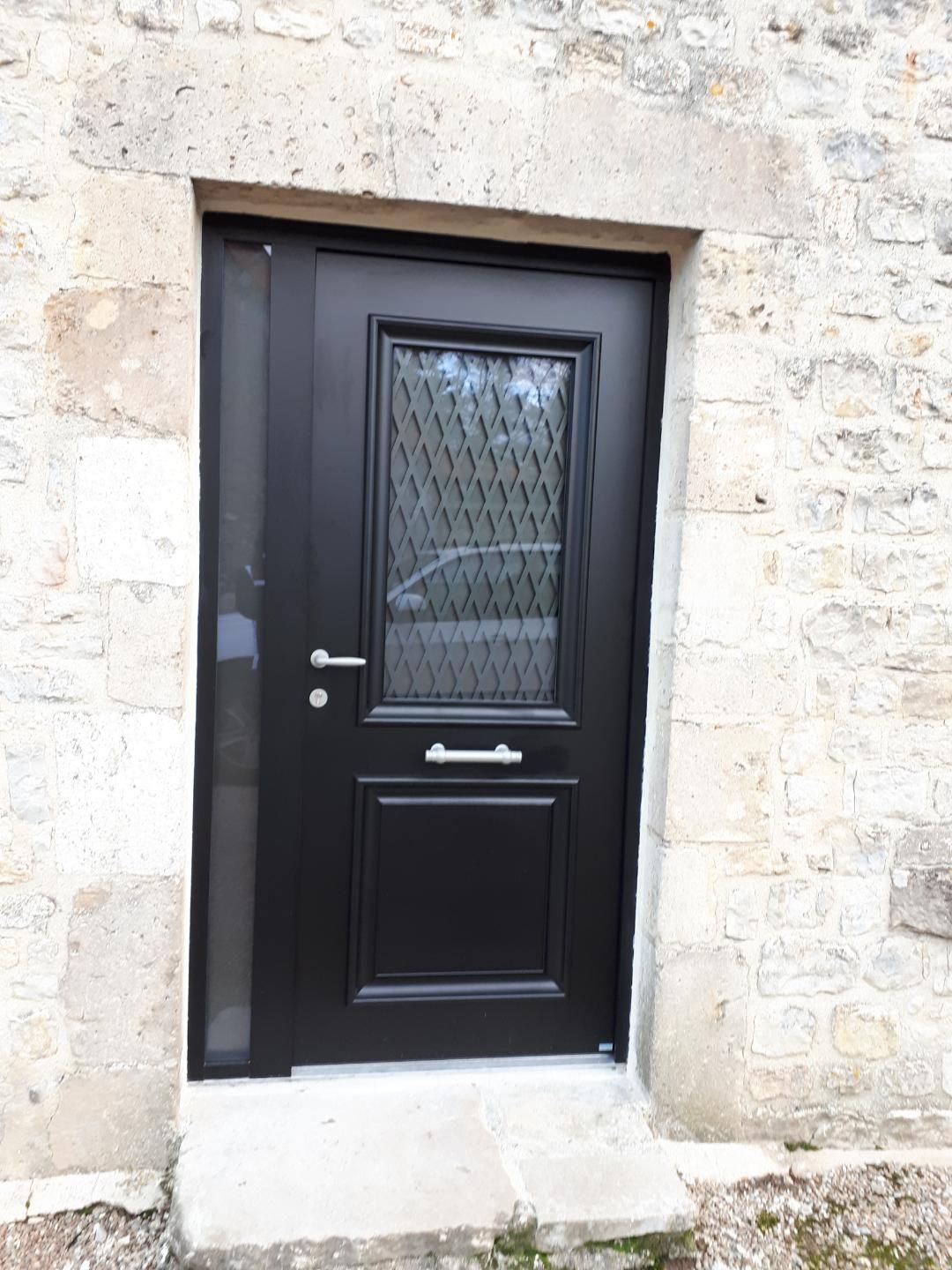 Porte d'entrée noire avec verre dépoli encastrée dans un mur en pierre, vue de l'extérieur.