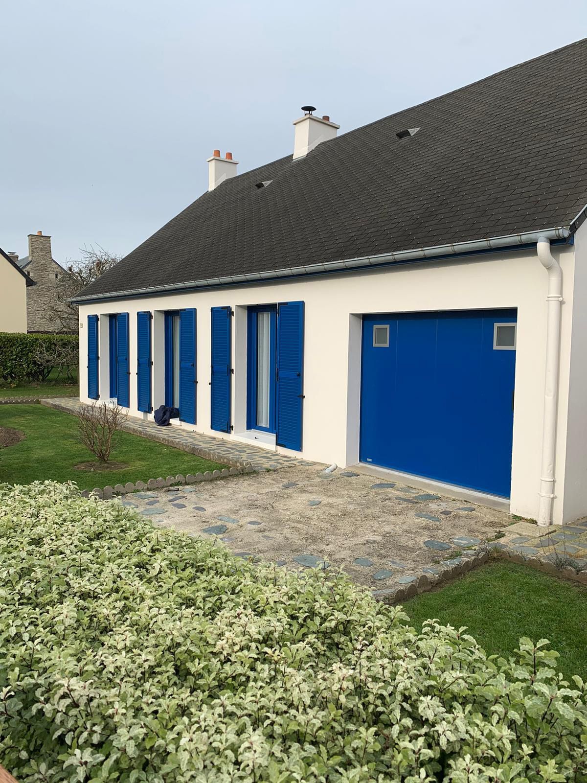 Maison blanche aux volets et portes bleus, bordée d'un chemin de gravier et de verdure basse.