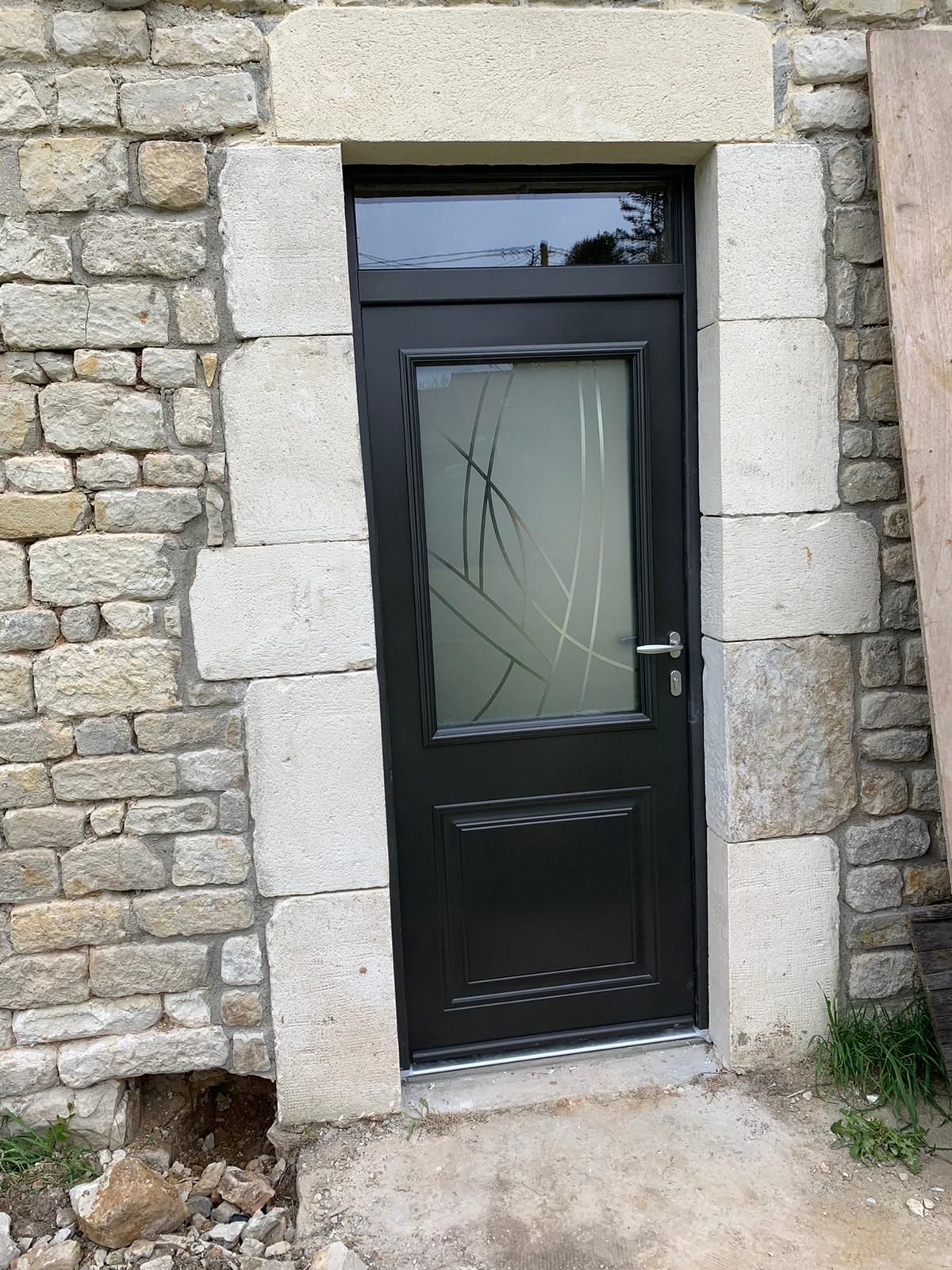 Porte d'entrée noire avec verre dépoli, encadrée par un mur en pierre.