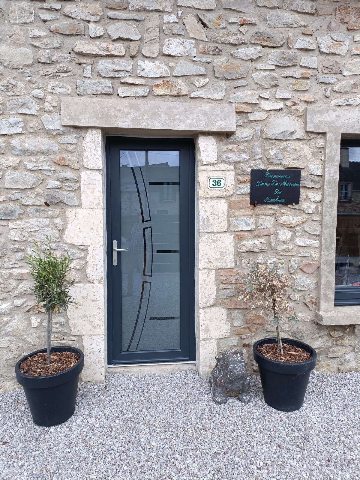 Entrée d'une maison en pierre avec une porte gris foncé, deux plantes en pot et un petit panneau à côté de la porte.