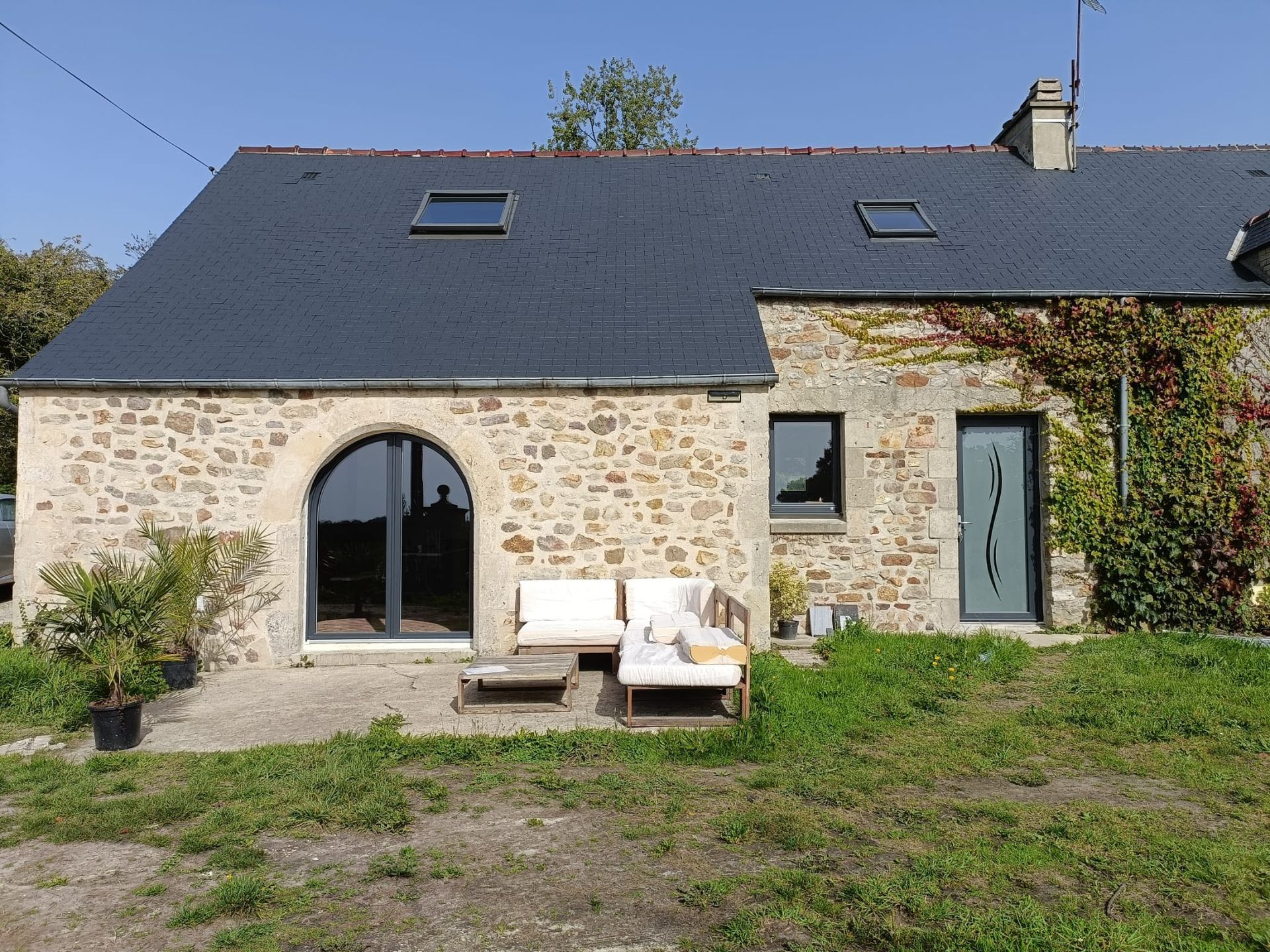Maisonnette en pierre avec toit sombre, porte vitrée cintrée et canapé d'extérieur dans une cour ensoleillée