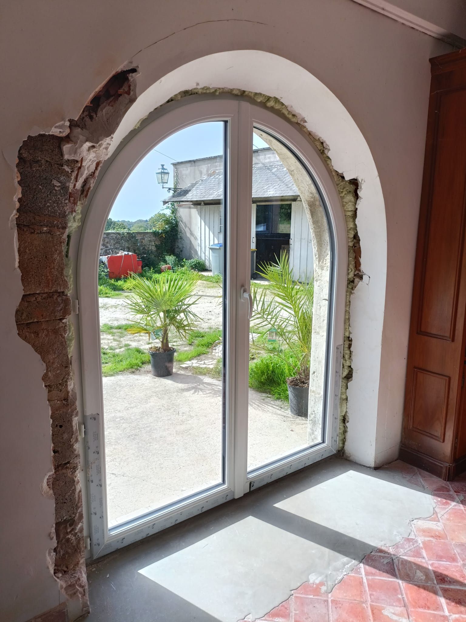 Portes-fenêtres cintrées en verre donnant sur un jardin ensoleillé, avec un enduit abîmé autour du cadre.