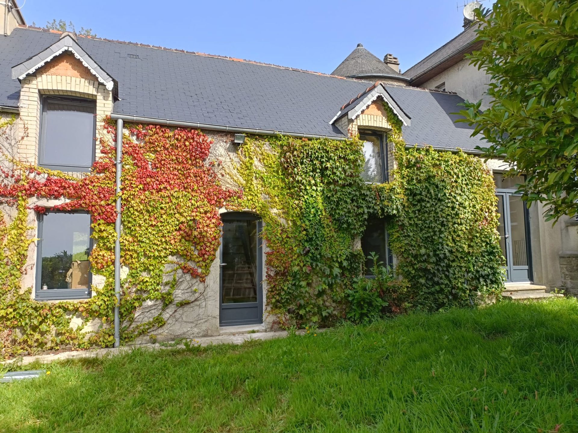 Maison en pierre recouverte de lierre rouge, vert et jaune, au bord d'une pelouse sous un ciel bleu.