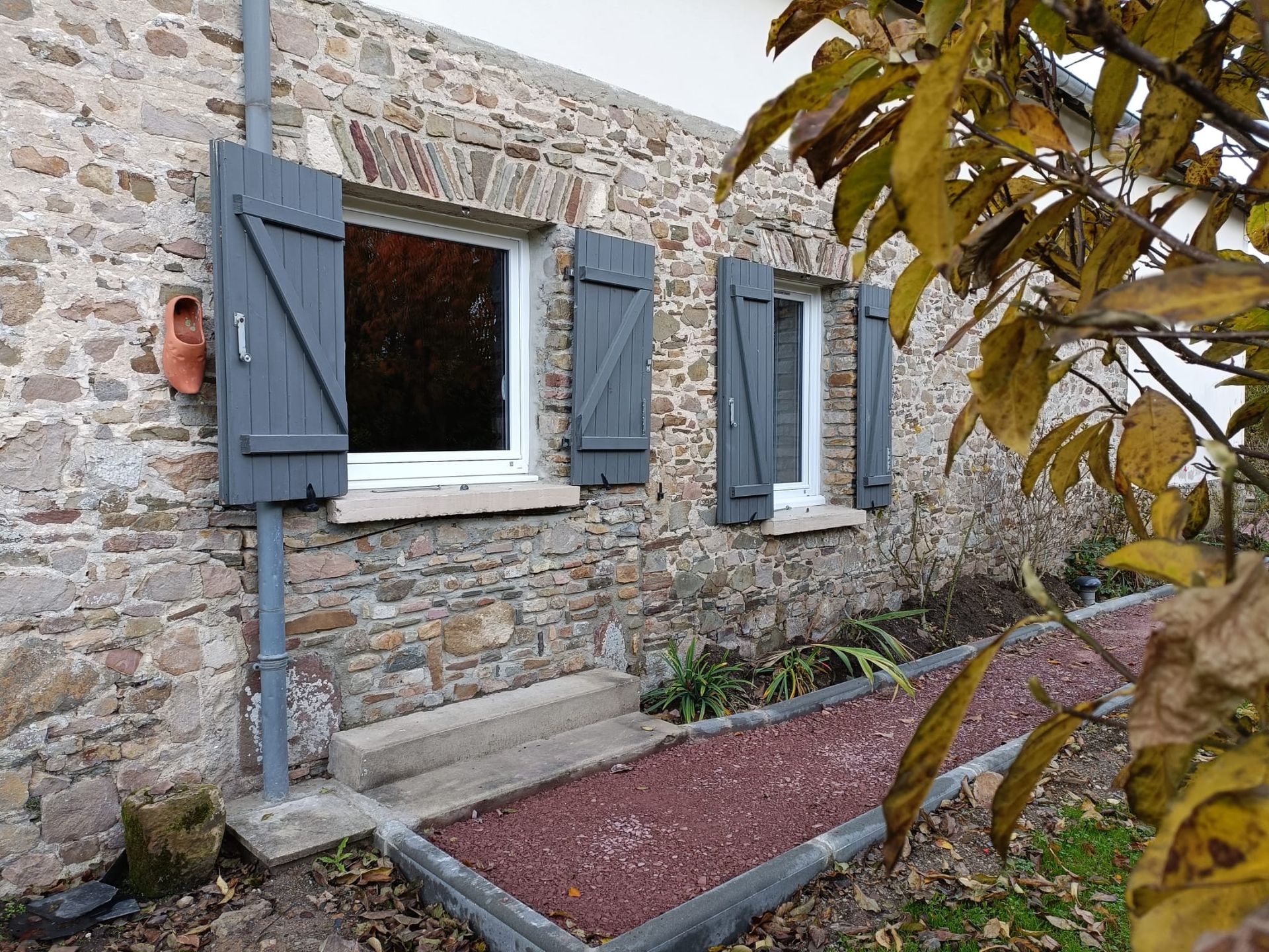 Mur en pierre d'une maison avec une fenêtre blanche et des volets gris, à côté d'un parterre de gravier et de branches d'arbres feuillues