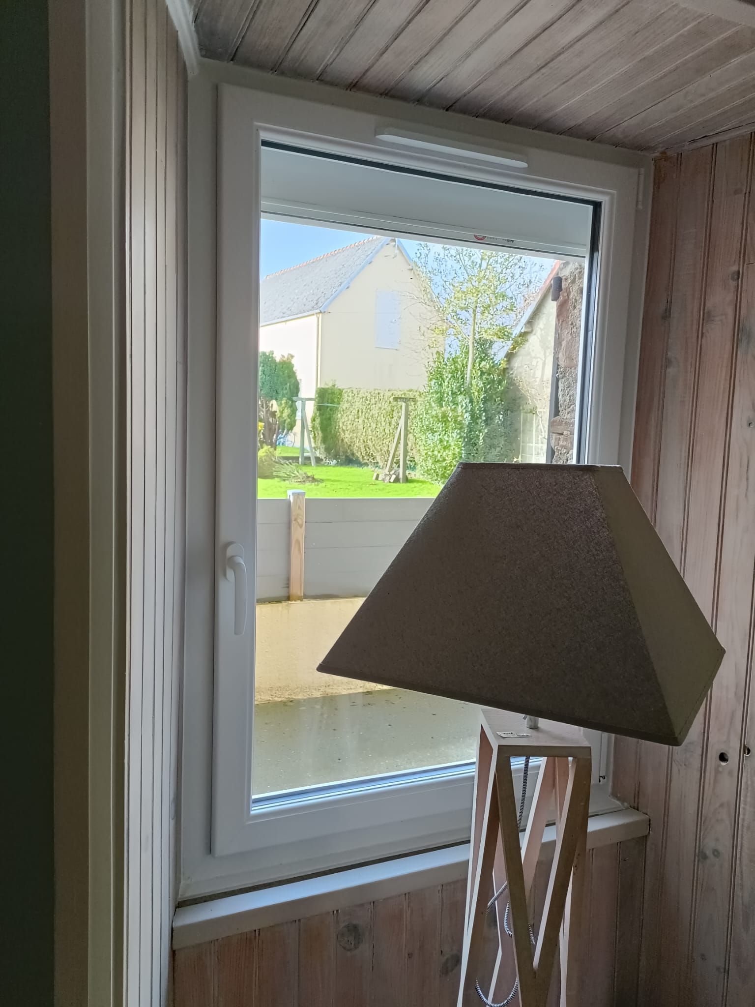 Vue depuis une fenêtre d'une pièce avec une lampe beige au premier plan, donnant sur une cour résidentielle ensoleillée à l'extérieur
