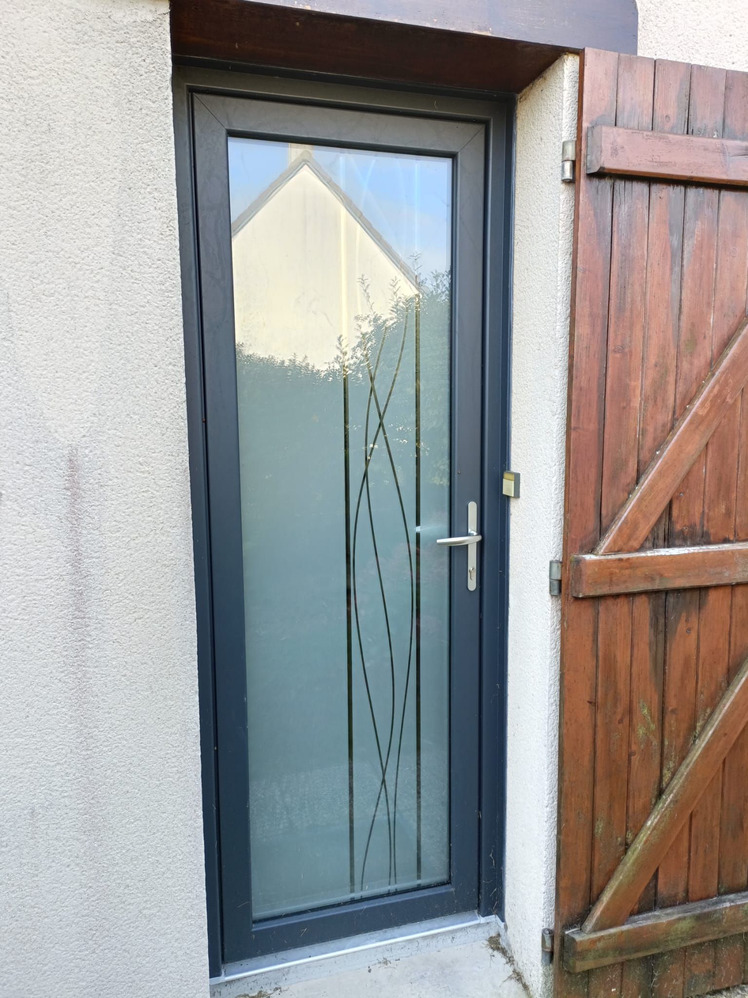 Porte latérale en verre dépoli avec poignée en métal dans un mur en stuc à côté d'un portail en bois