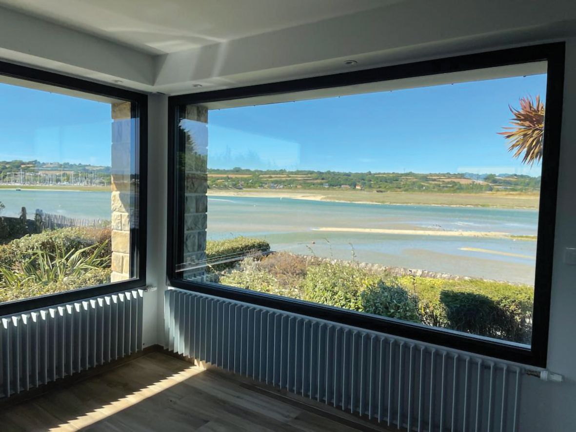 Chambre d'angle avec de grandes fenêtres donnant sur une baie calme et le rivage au loin, sous un ciel bleu azur.