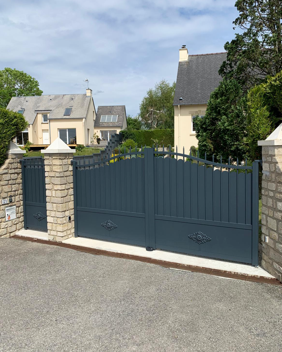 Un portail bleu foncé entre des piliers en pierre, devant des maisons beiges dans une rue ensoleillée