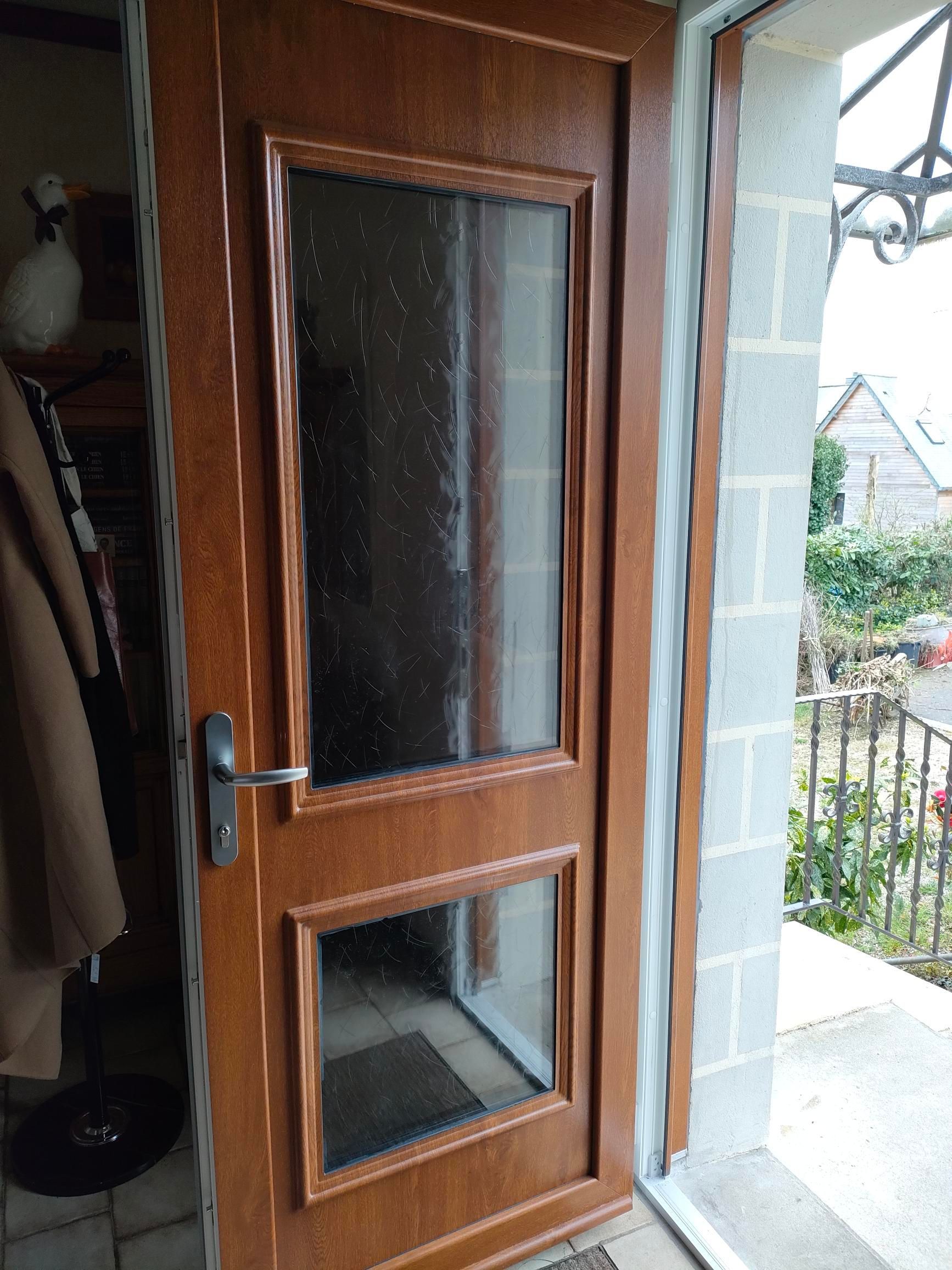 La porte d'entrée en bois et vitrée est légèrement ouverte, laissant entrevoir l'entrée et le jardin extérieur.
