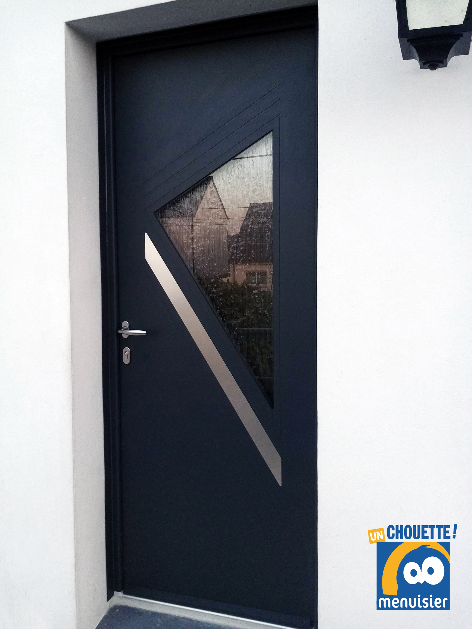 Porte d'entrée sombre avec une fenêtre triangulaire en verre et une bande diagonale dépolie sur un mur extérieur blanc.