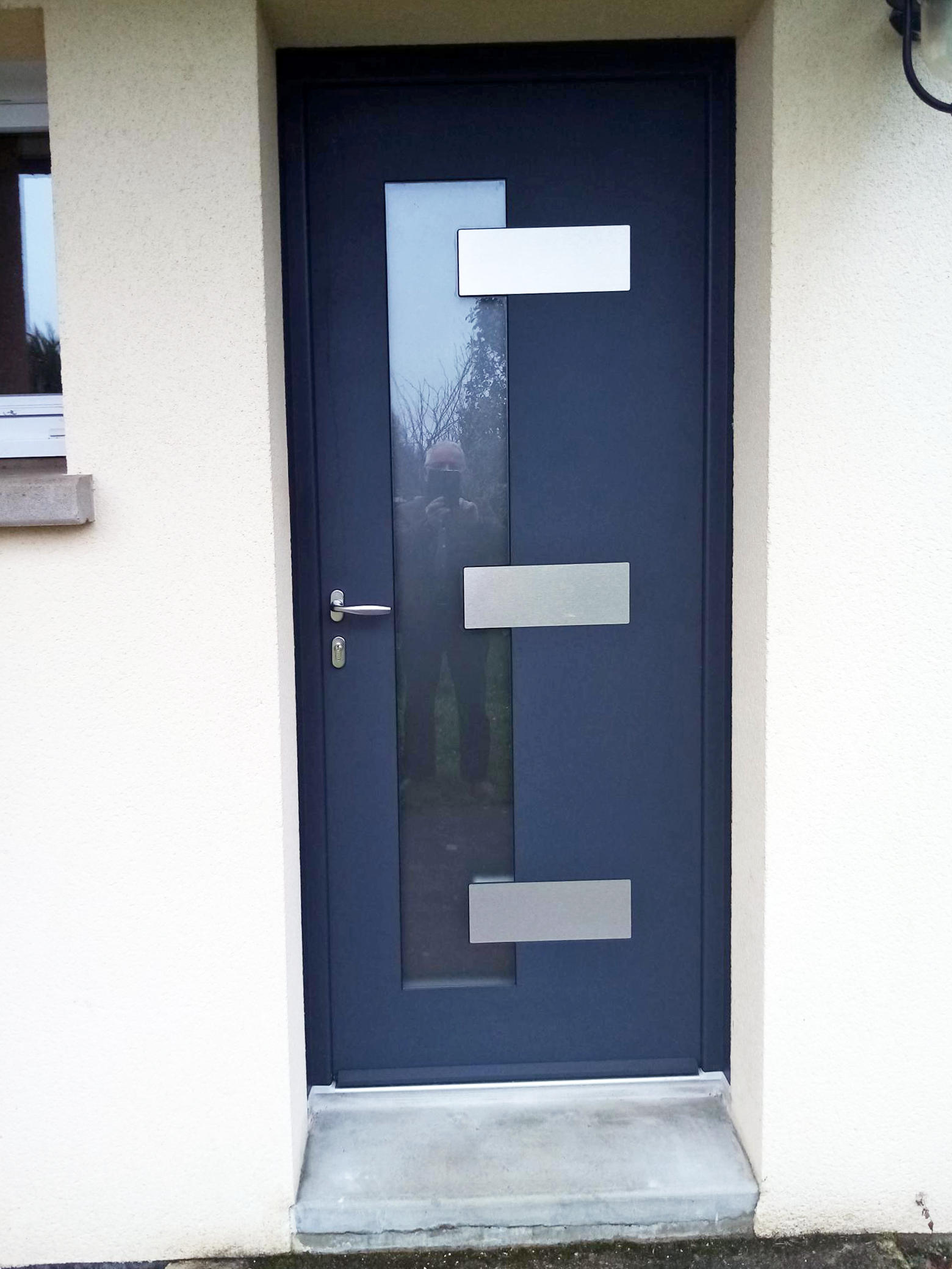 Porte d'entrée bleu foncé avec trois panneaux de verre dépoli et une poignée argentée, sur un mur extérieur beige.