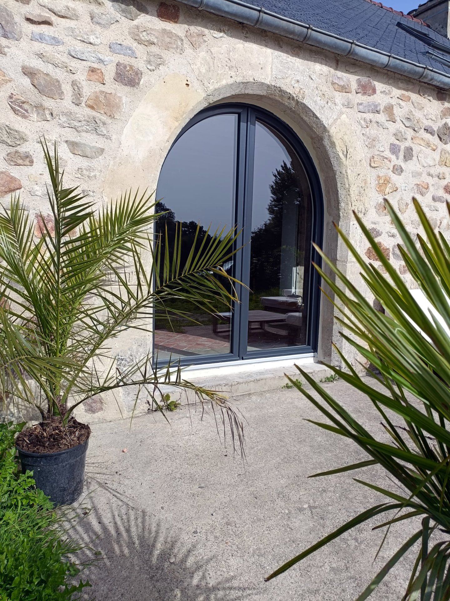 Porte cintrée en pierre avec porte vitrée à cadre noir, encadrée de palmiers en pot sur une terrasse en gravier.