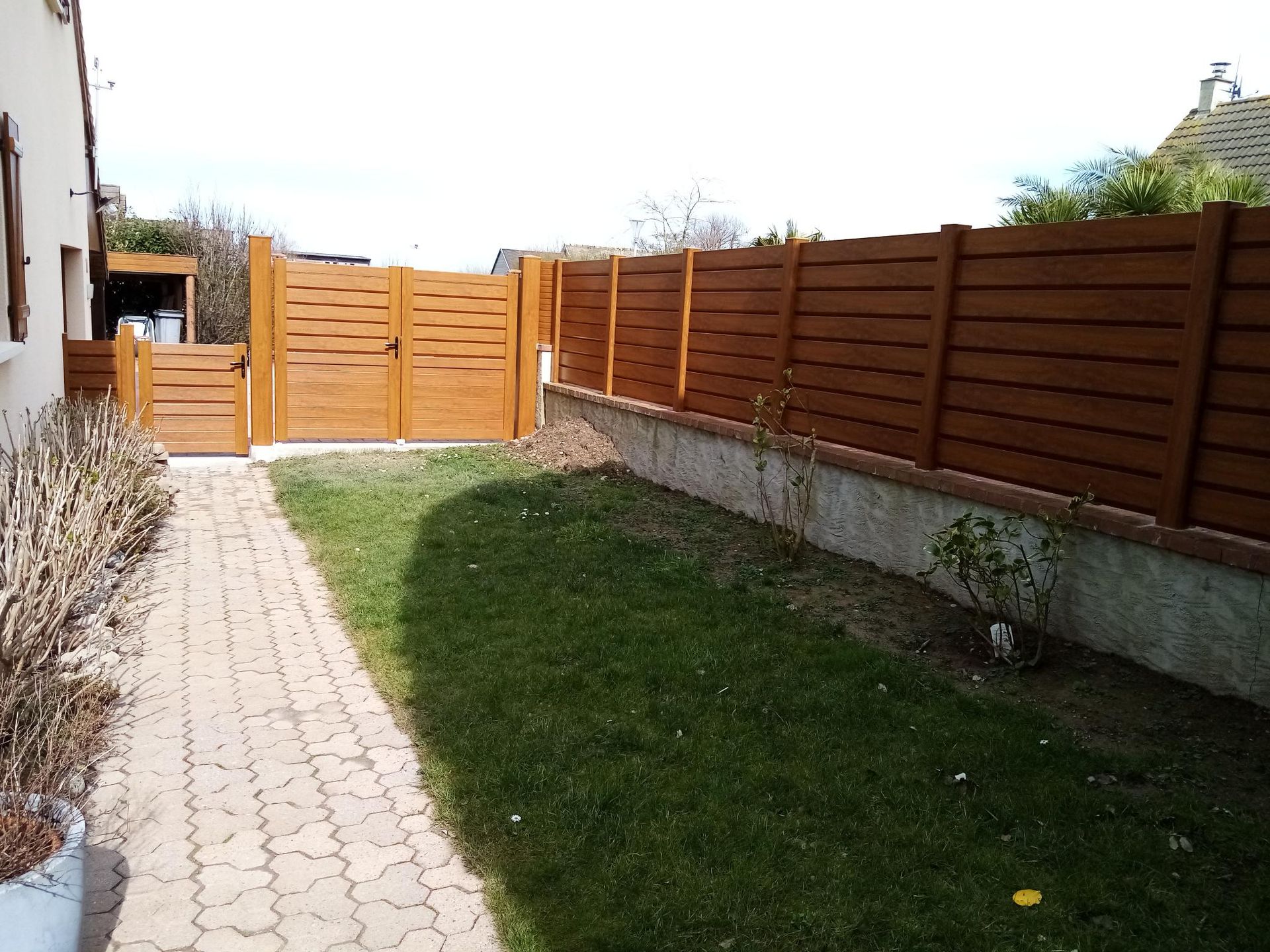 Cour arrière étroite avec pelouse, allée pavée, clôture en bois et portail au loin.