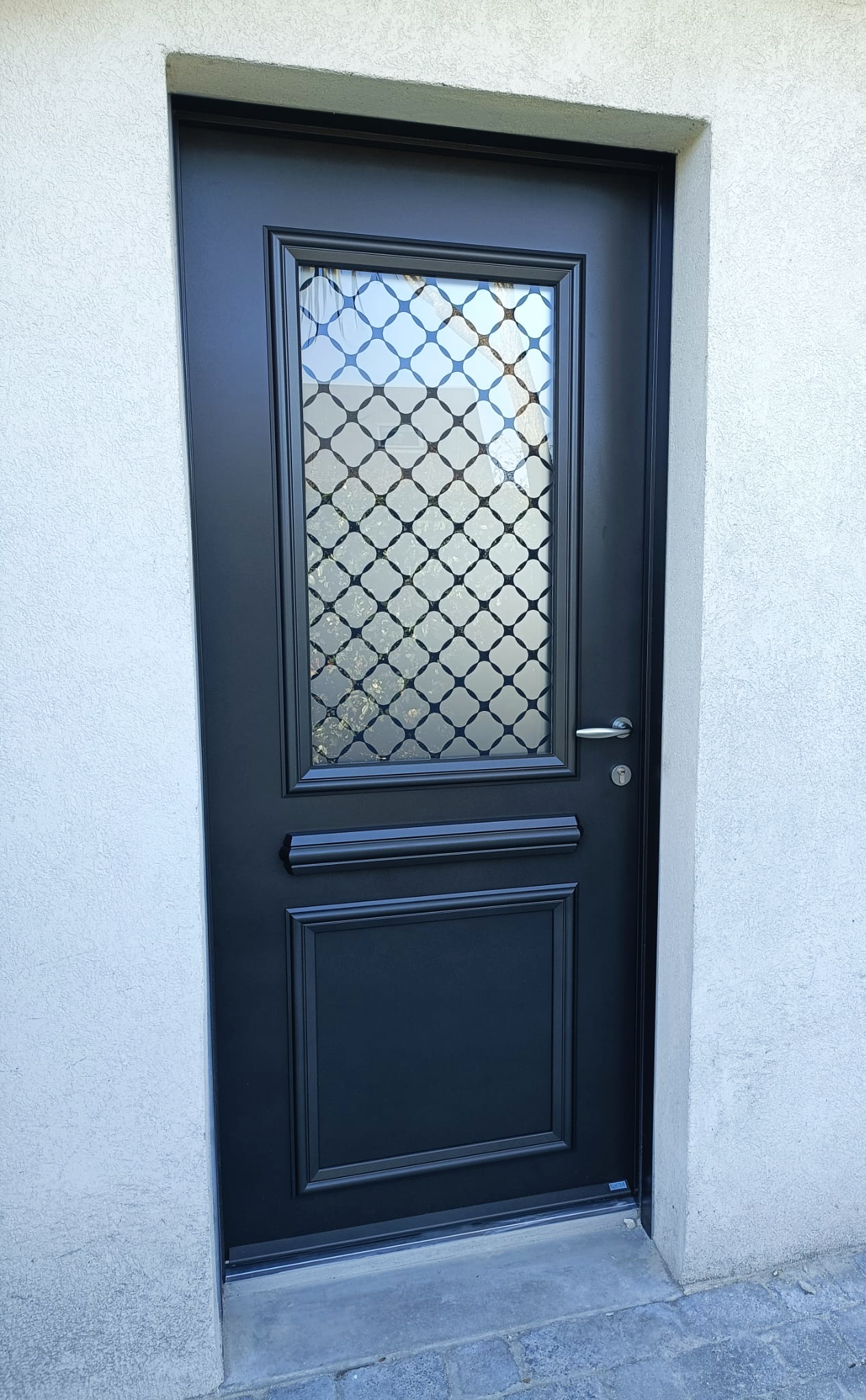 Porte extérieure noire avec un panneau vitré décoratif et une grille de sécurité, dans une entrée murale en stuc
