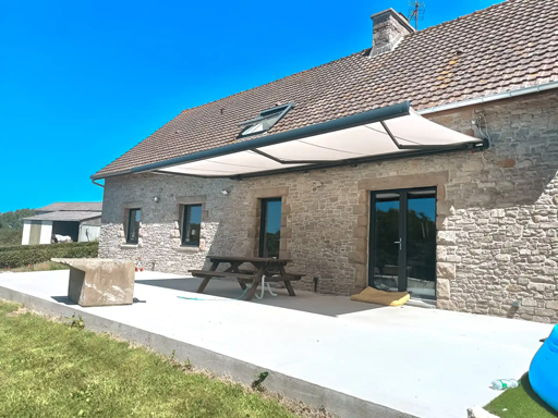 Terrasse en pierre avec auvent blanc, table de pique-nique et jardin gazonné sous un ciel bleu