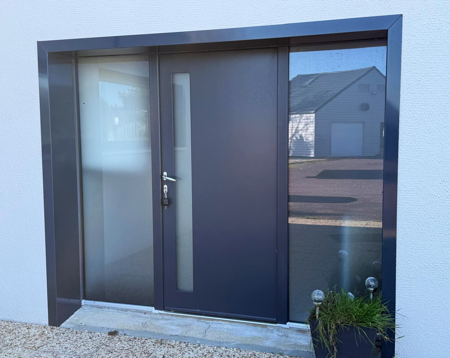 Porte d’entrée moderne avec panneau latéral en verre dépoli et reflet dans la fenêtre sur un mur extérieur en stuc
