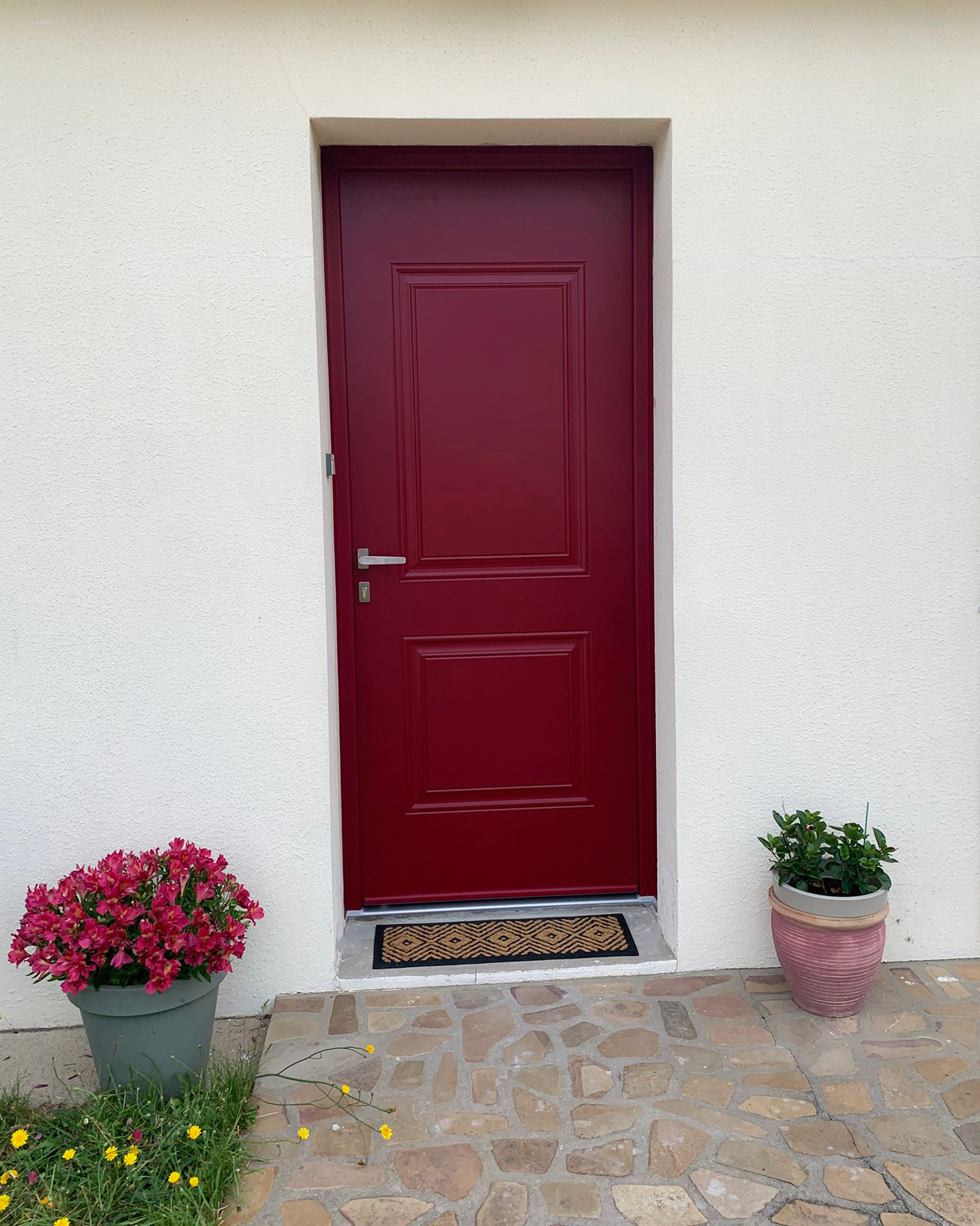 Porte d’entrée rouge avec des fleurs en pot de chaque côté, dans un mur en stuc clair.