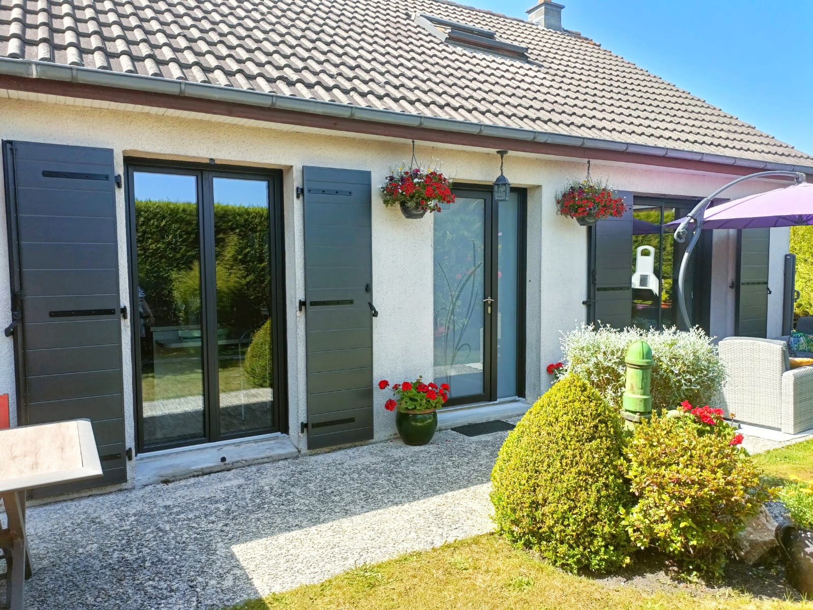 Façade de maison blanche baignée de soleil, avec des volets noirs, des pots de fleurs et un patio de jardin en gravier