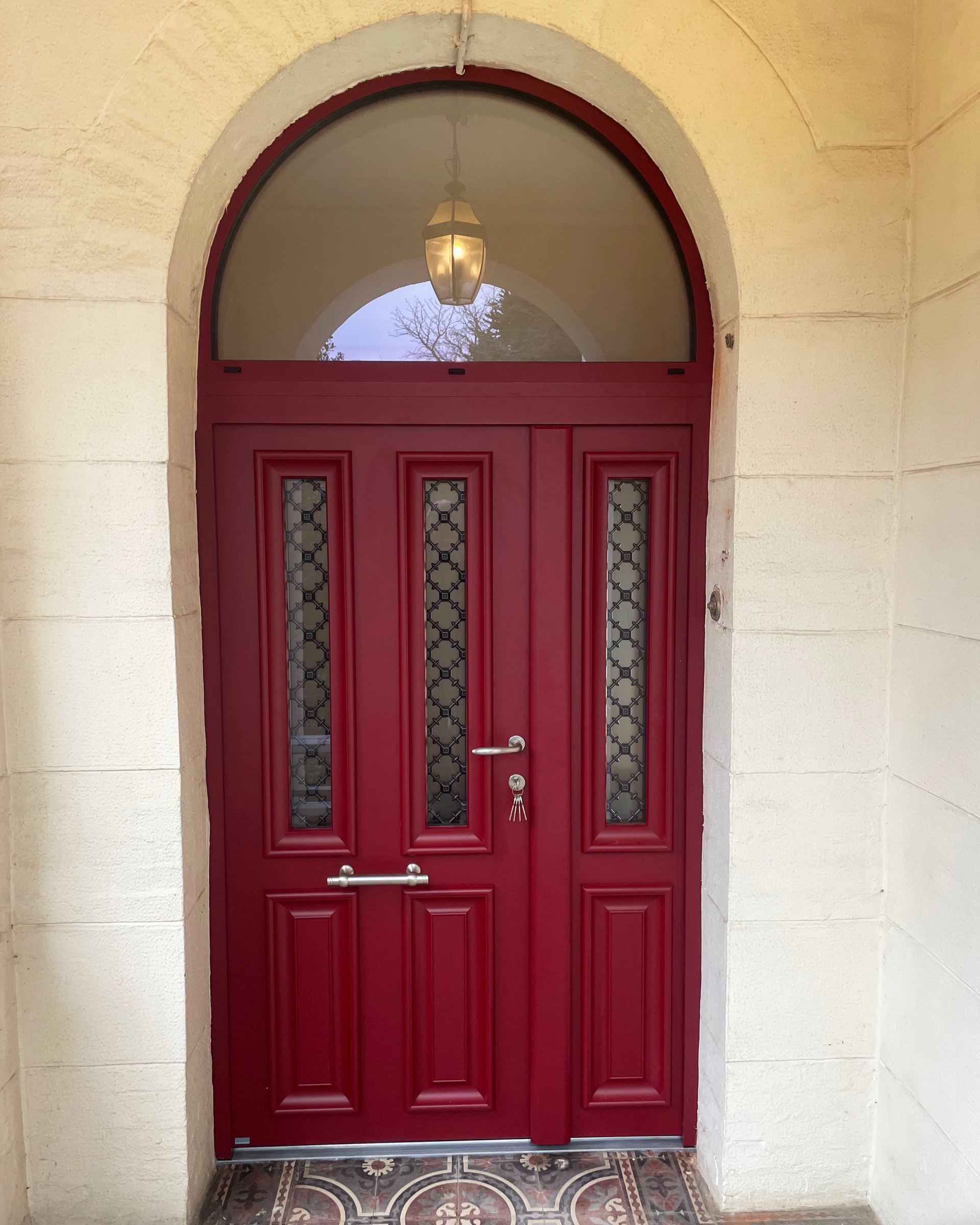 Double porte d’entrée rouge avec fenêtre en arc dans un mur en stuc beige.
