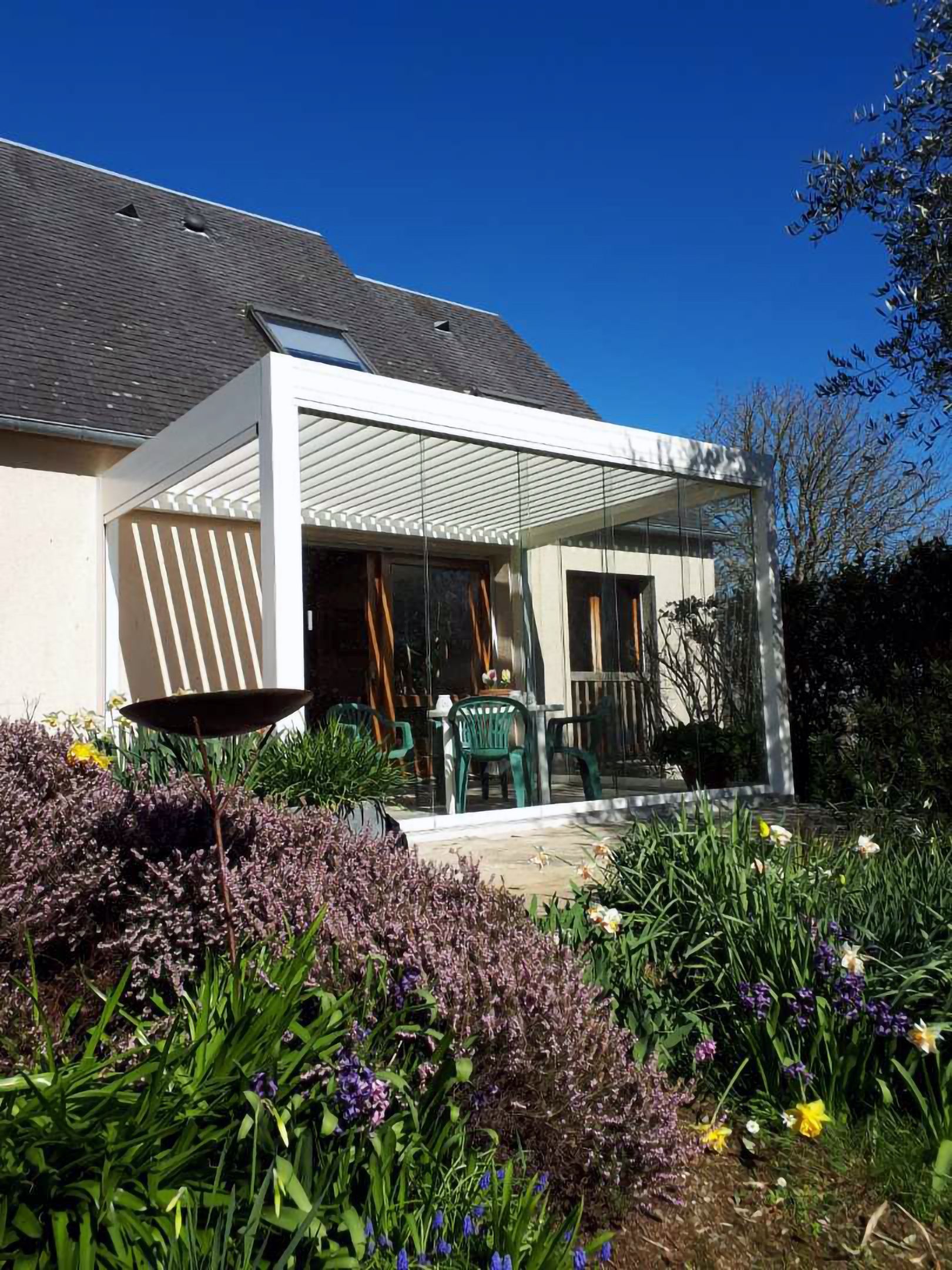 Patio d'une maison avec pergola blanche, chaises vertes et fleurs violettes au premier plan sous un ciel bleu