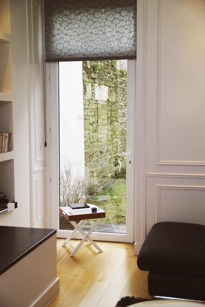 Chambre moderne et lumineuse avec porte-fenêtre donnant sur un balcon, petite table près des plantes et canapé foncé contre le mur.