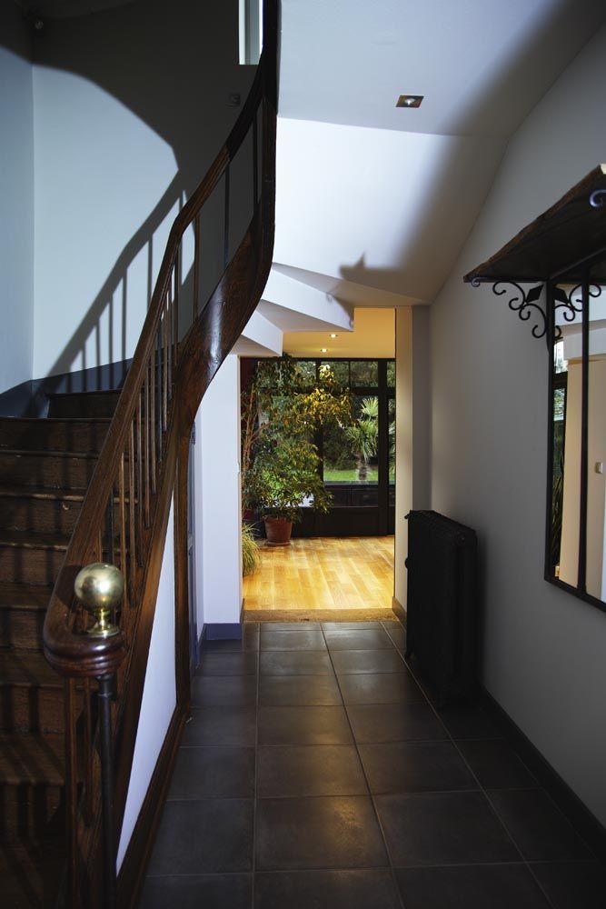 Couloir étroit au sol carrelé foncé, escalier en colimaçon, miroir et lumière naturelle donnant sur une terrasse de jardin