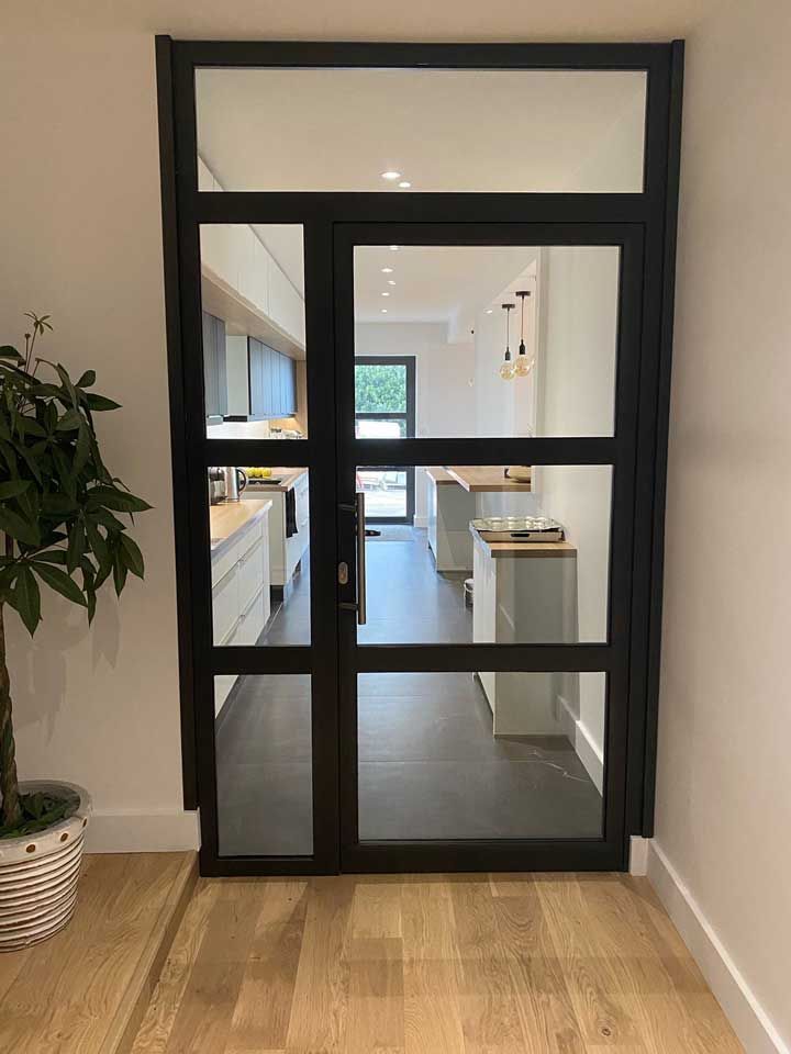 Une porte vitrée à cadre noir s'ouvre sur une cuisine lumineuse avec un parquet clair et des meubles blancs.