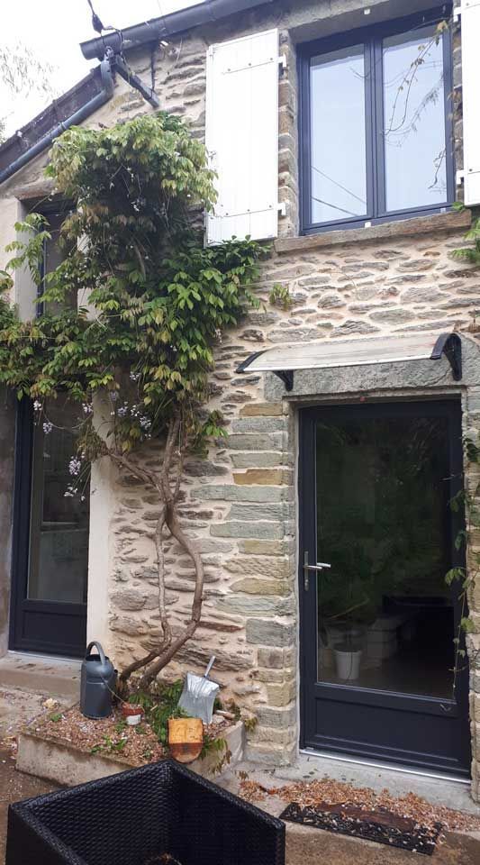 Maison en pierre avec porte noire, volets blancs et lierre grimpant près d'un petit arbre