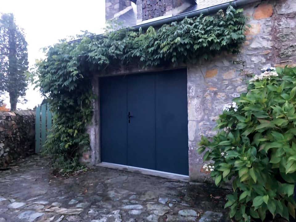 Double porte bleue dans un mur de pierre, encadrée de lierre et de verdure, sur un chemin pavé.