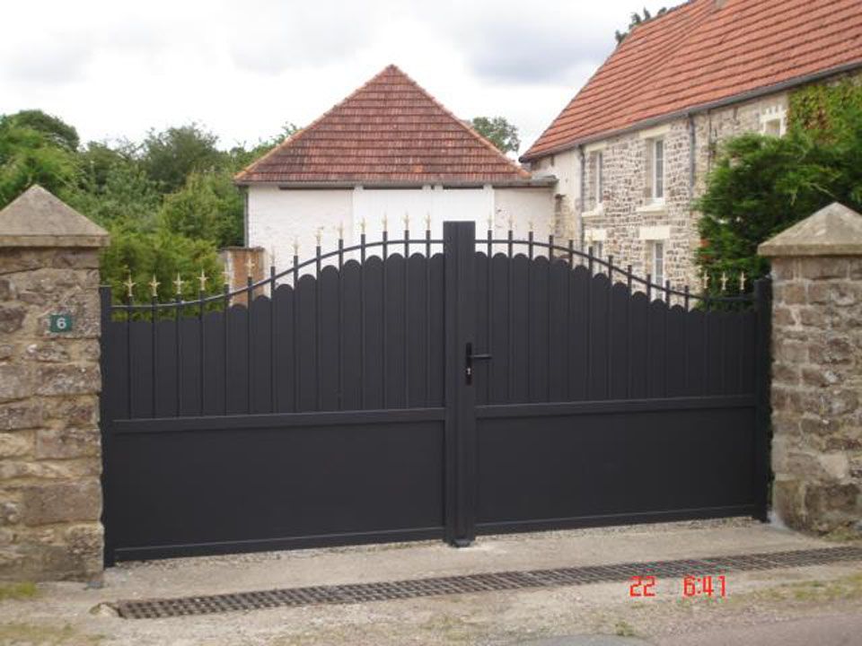 Grand portail d'allée en métal noir entre des piliers de pierre devant une maison aux toits de tuiles rouges.