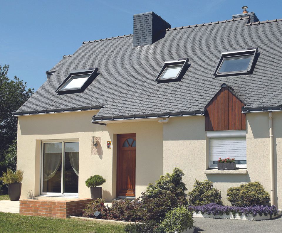 Maison couleur crème avec toit en ardoise foncée, puits de lumière et jardin à l'avant sous un ciel bleu azur.