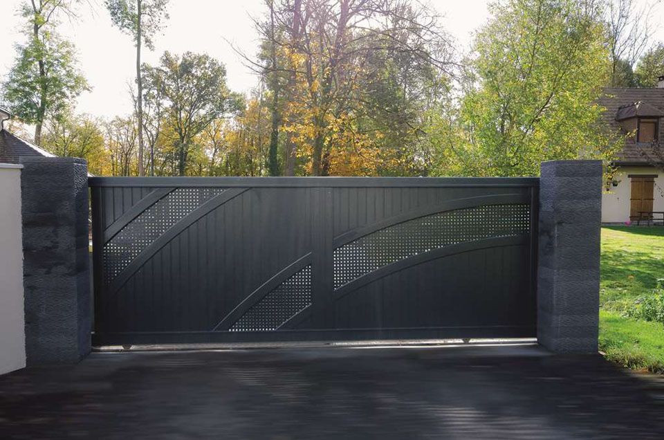 Portail d'allée en métal sombre entre des piliers de pierre, avec des arbres et une maison en arrière-plan.