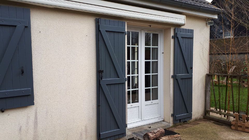 Portes-fenêtres à volets bleus sur une façade beige, à côté d'une petite terrasse et d'une clôture de jardin