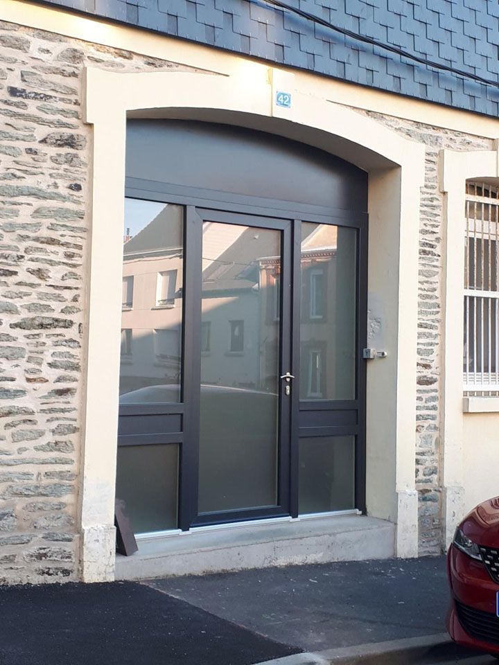 Entrée d'un bâtiment en pierre avec des portes doubles en verre foncé et un trottoir étroit devant.