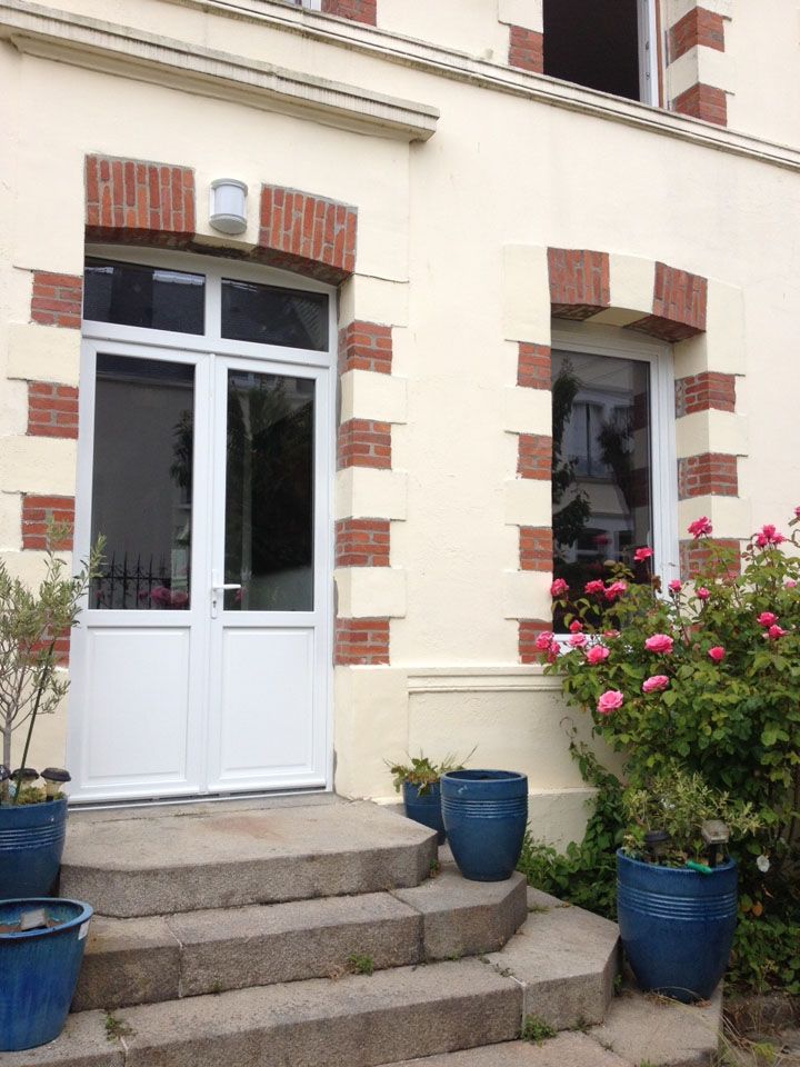Porte d'entrée d'une maison couleur crème avec marches en pierre et pots de fleurs bleus, flanquée de buissons à fleurs roses.
