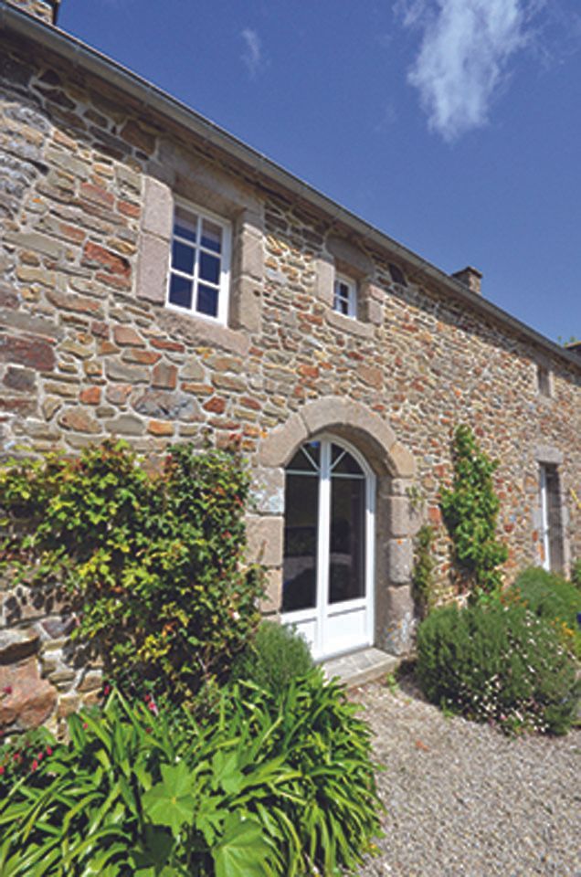 Extérieur d'un cottage en pierre avec une porte cintrée blanche, des arbustes verts et un chemin de gravier sous un ciel bleu.