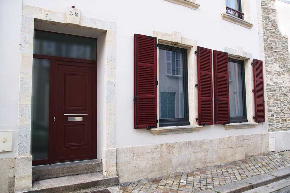 Porte rouge et fenêtres à volets sur une façade de maison en pierre blanche