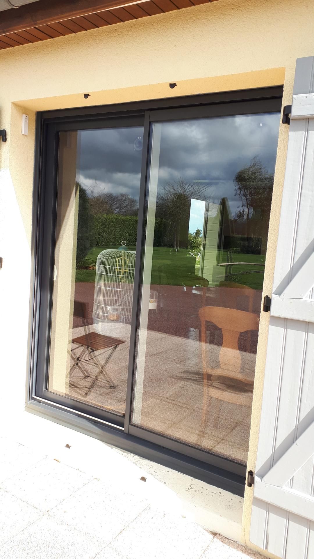 Porte coulissante en verre à cadre noir et volets blancs reflétant une terrasse et un jardin verdoyant.