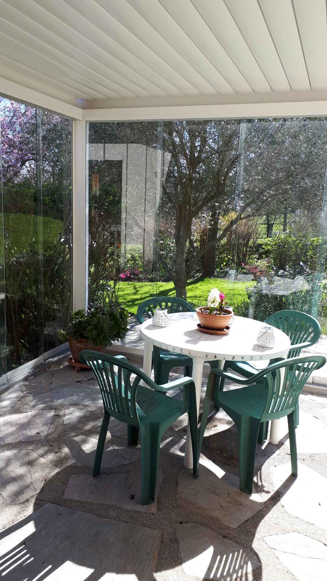 Terrasse couverte avec des chaises vertes autour d'une table blanche dans un jardin ensoleillé.