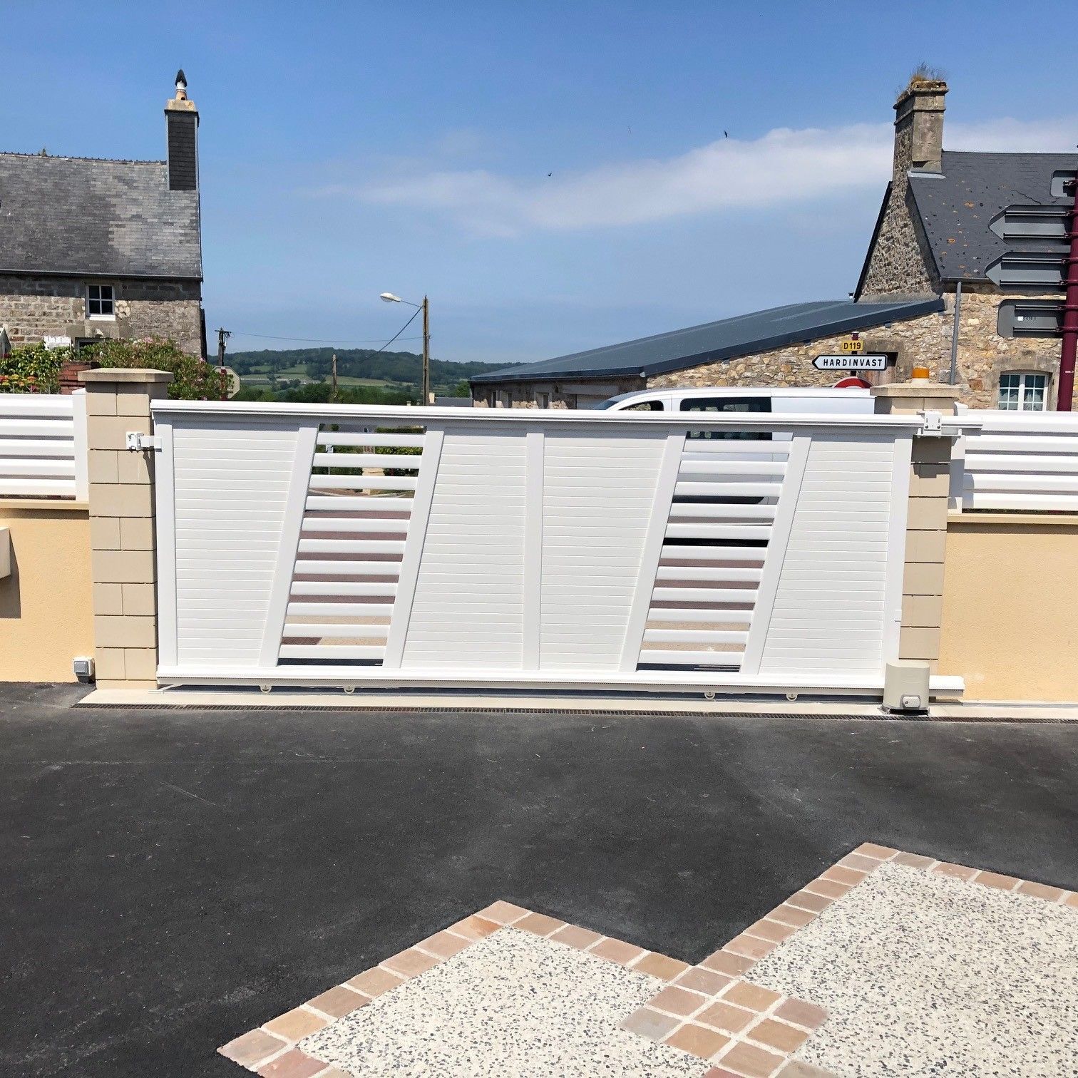 Portail coulissant blanc sur un mur couleur crème, avec des maisons en pierre et une colline au loin sous un ciel bleu.