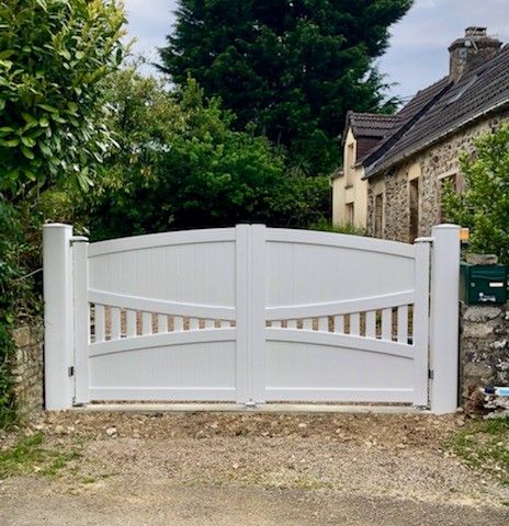 Portail d'allée en bois blanc devant une maison en pierre et de la verdure.